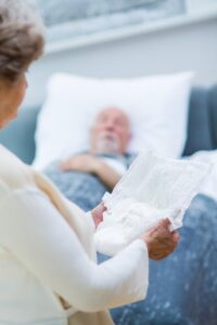 Ältere Frau steht vor dem Bett ihres kranken Mannes und hält eine Windel für Erwachsene